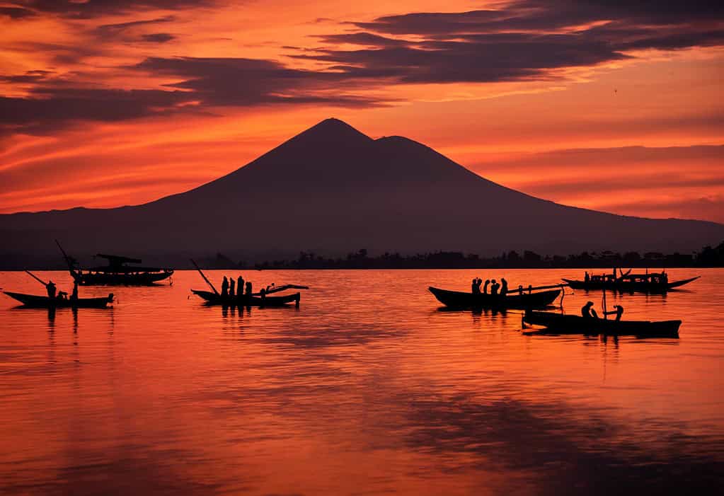 Silhouettes of fishermen in traditional wooden boats against a fiery sky reflecting in the calm waters with Nyiragongo volcano in the background